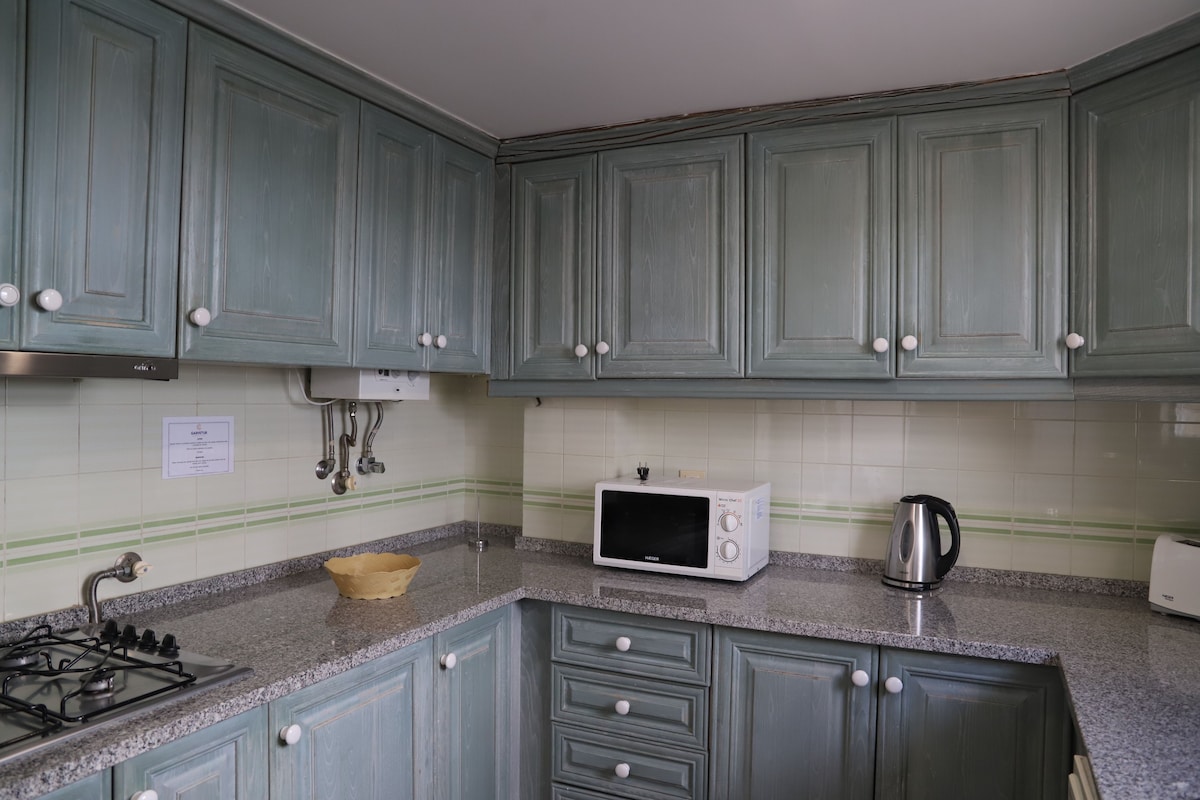 Een keuken met blauwgroene kastjes, een magnetron, een waterkoker en een gasfornuis.
