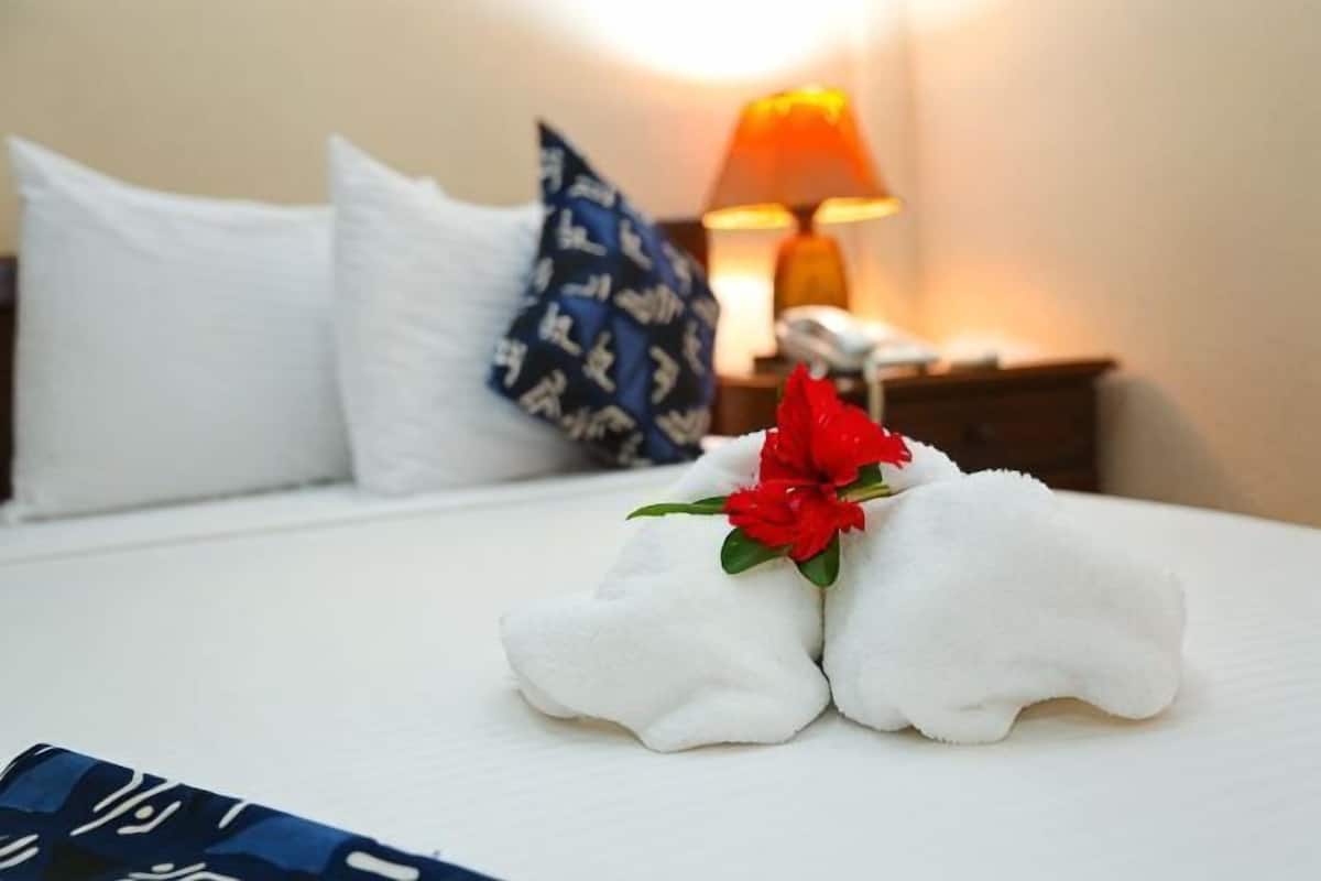 Una cama bien hecha con una toalla y una flor roja, almohadas blancas y una lámpara al fondo.