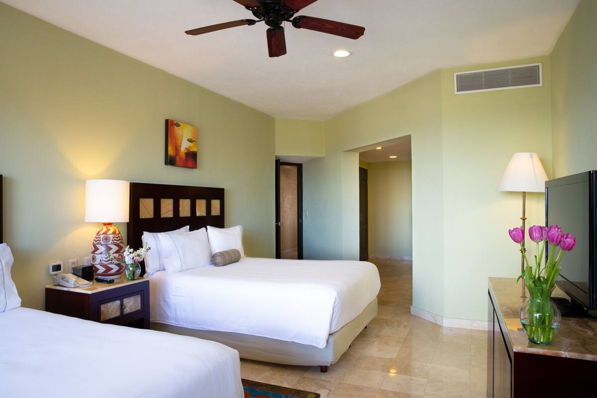 A hotel room with two beds, a ceiling fan, a painting, a lamp, and a vase with flowers.