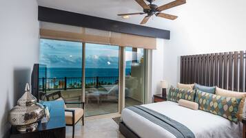 A modern hotel room with a large bed, a ceiling fan, a balcony with a view of the ocean, and a desk with a lamp.