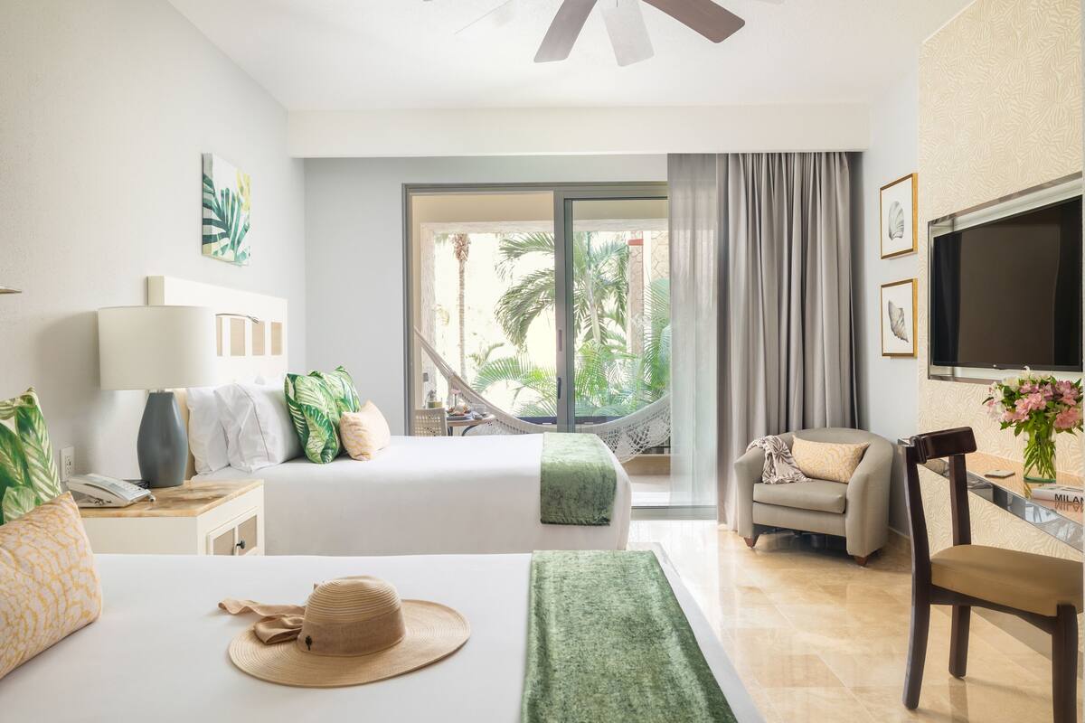 A hotel room with a bed, a desk, a chair, a TV, and a view of palm trees.