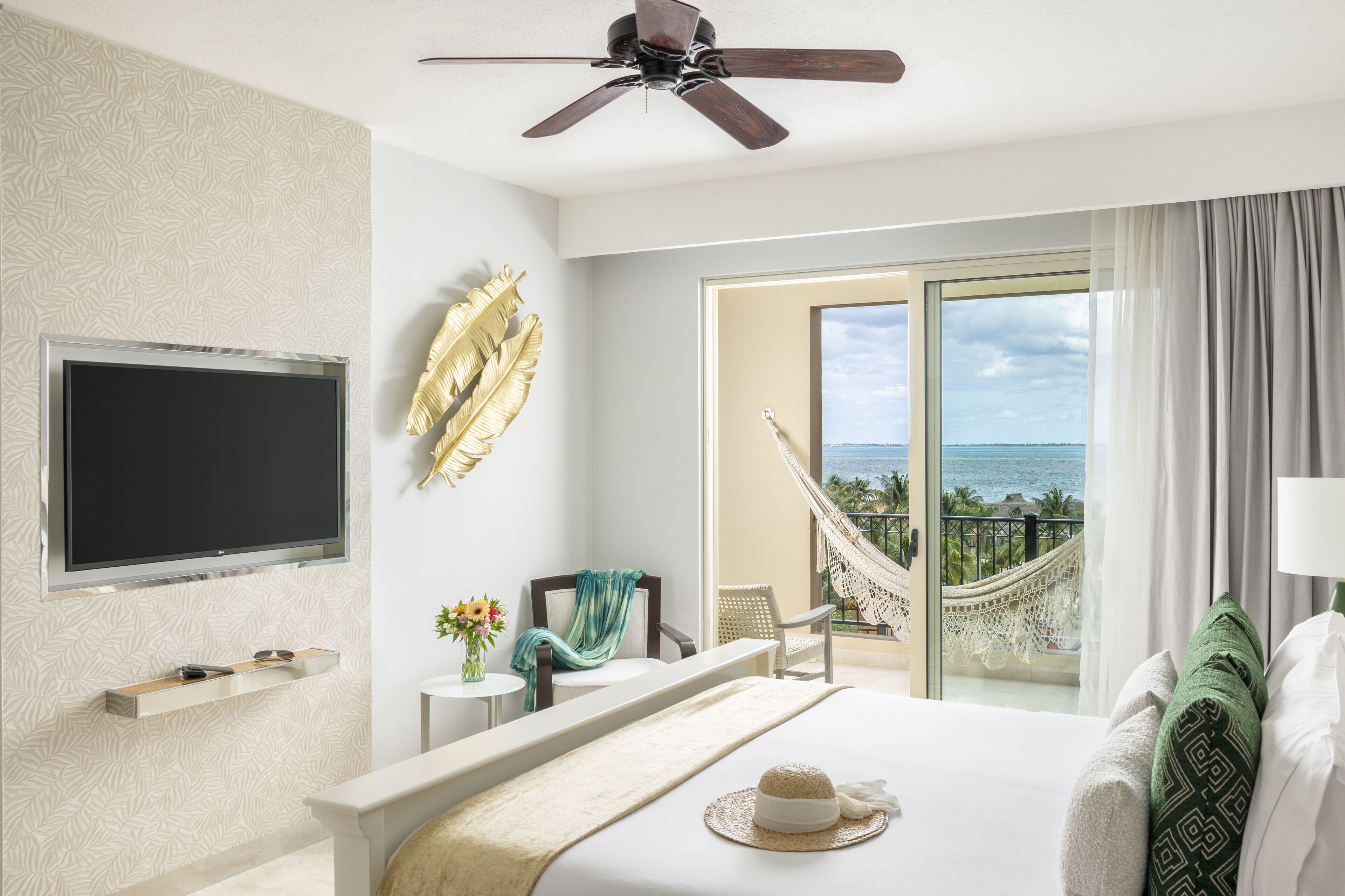 A hotel room with a bed, a TV, a hammock, a balcony with a view of the ocean, and a ceiling fan.