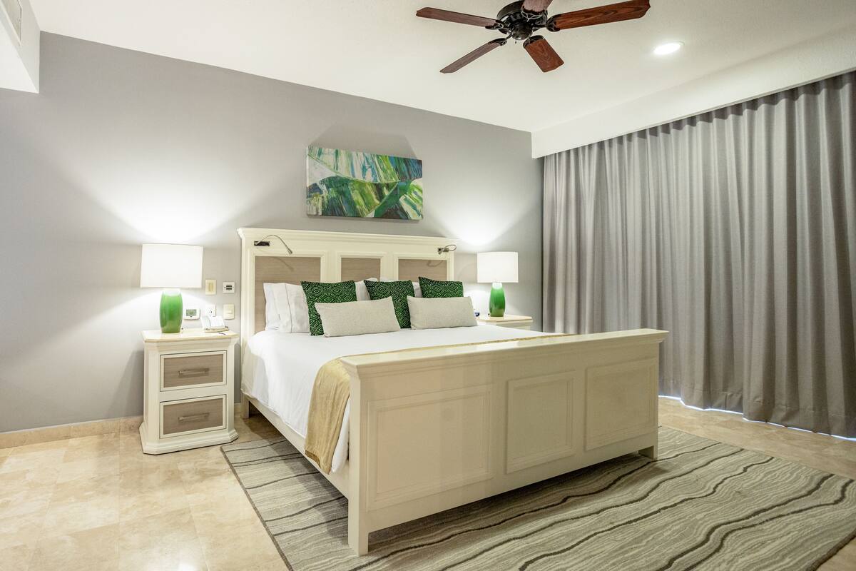 A bedroom with a bed, nightstands, a ceiling fan, a painting, and a window with curtains.