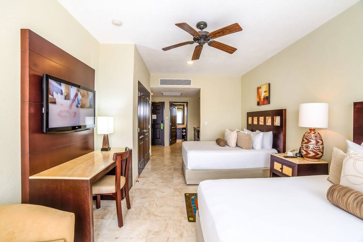 A hotel room with a TV, desk, two beds, a ceiling fan, and a hallway leading to a bathroom.