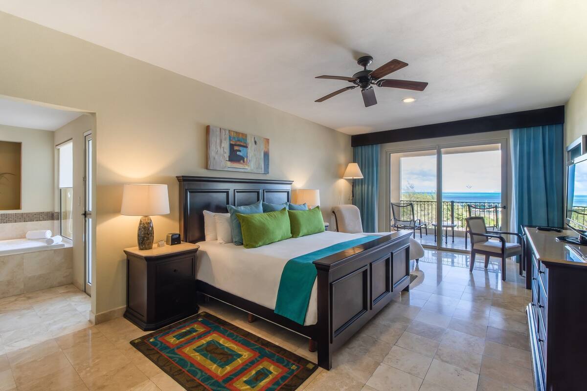 A hotel room with a bed, two bedside tables, a desk, a chair, a balcony with a view, and a ceiling fan.