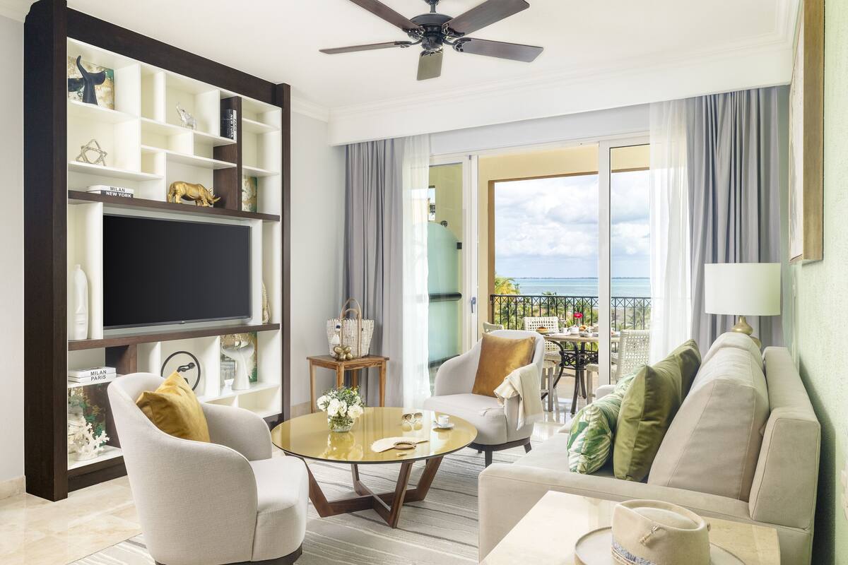 A modern living room with a flat-screen TV, comfortable seating, a balcony with a view, and a ceiling fan.