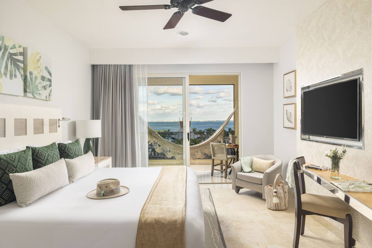 A hotel room with a bed, a desk, a chair, a TV, and a balcony with a view of the ocean.