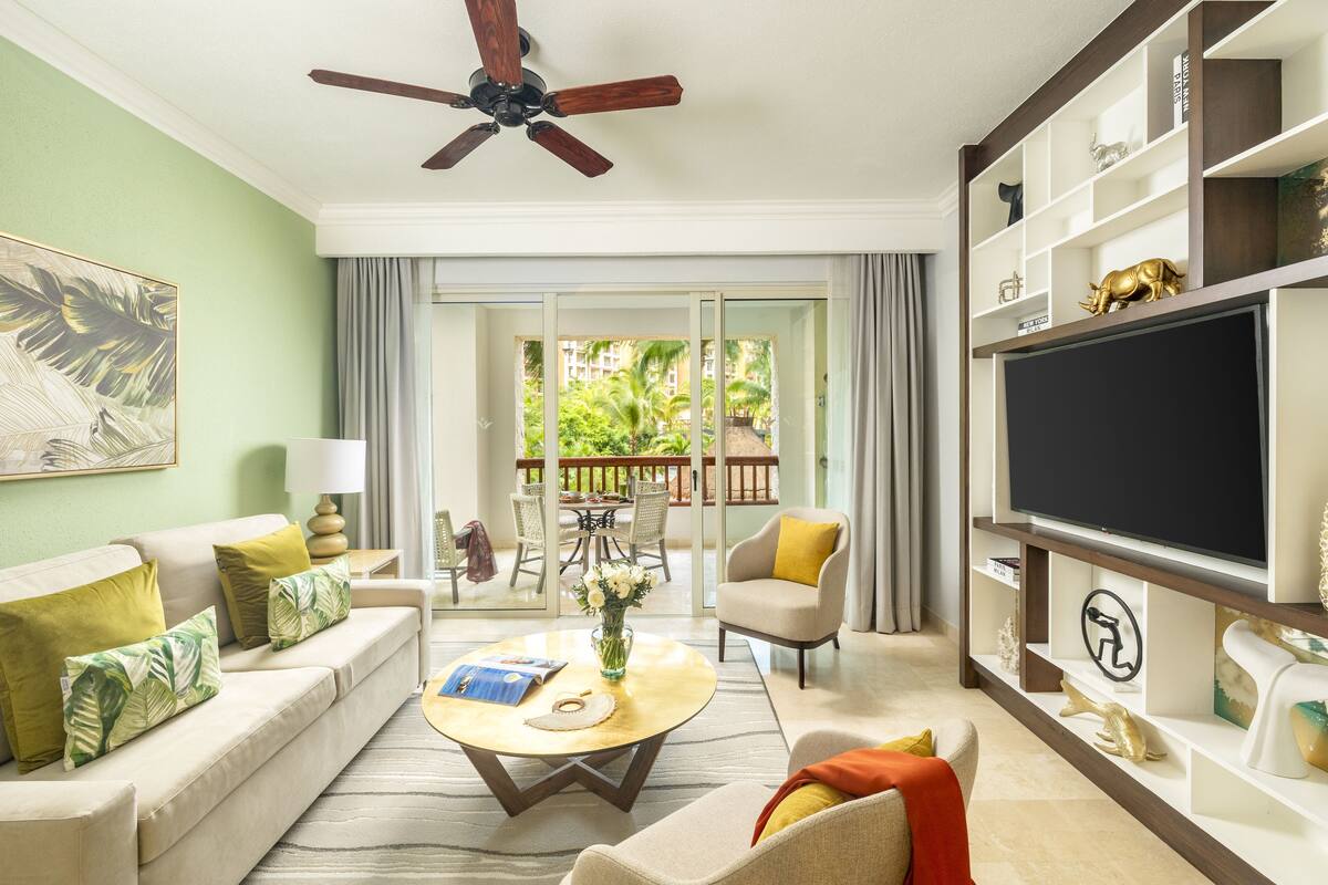 A living room with a sofa, armchair, coffee table, TV, and a balcony with outdoor furniture.