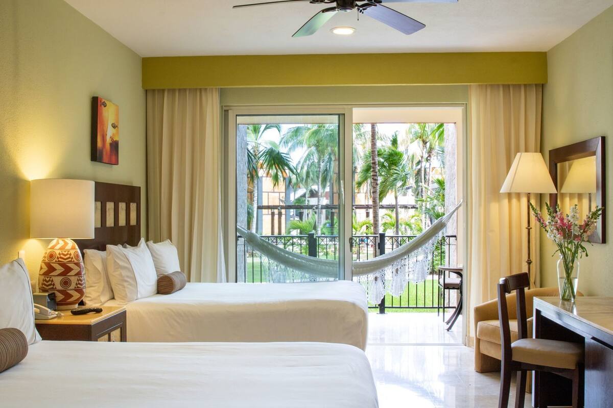A hotel room with two beds, a desk, a chair, a hammock, and a balcony with a view of palm trees.