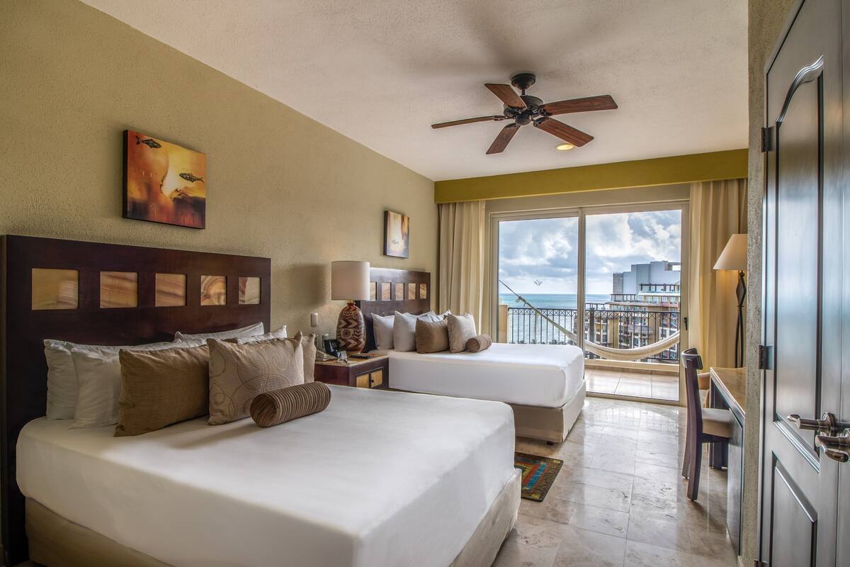 A hotel room with two beds, a ceiling fan, a large window with a view of the city, and a balcony with a railing.