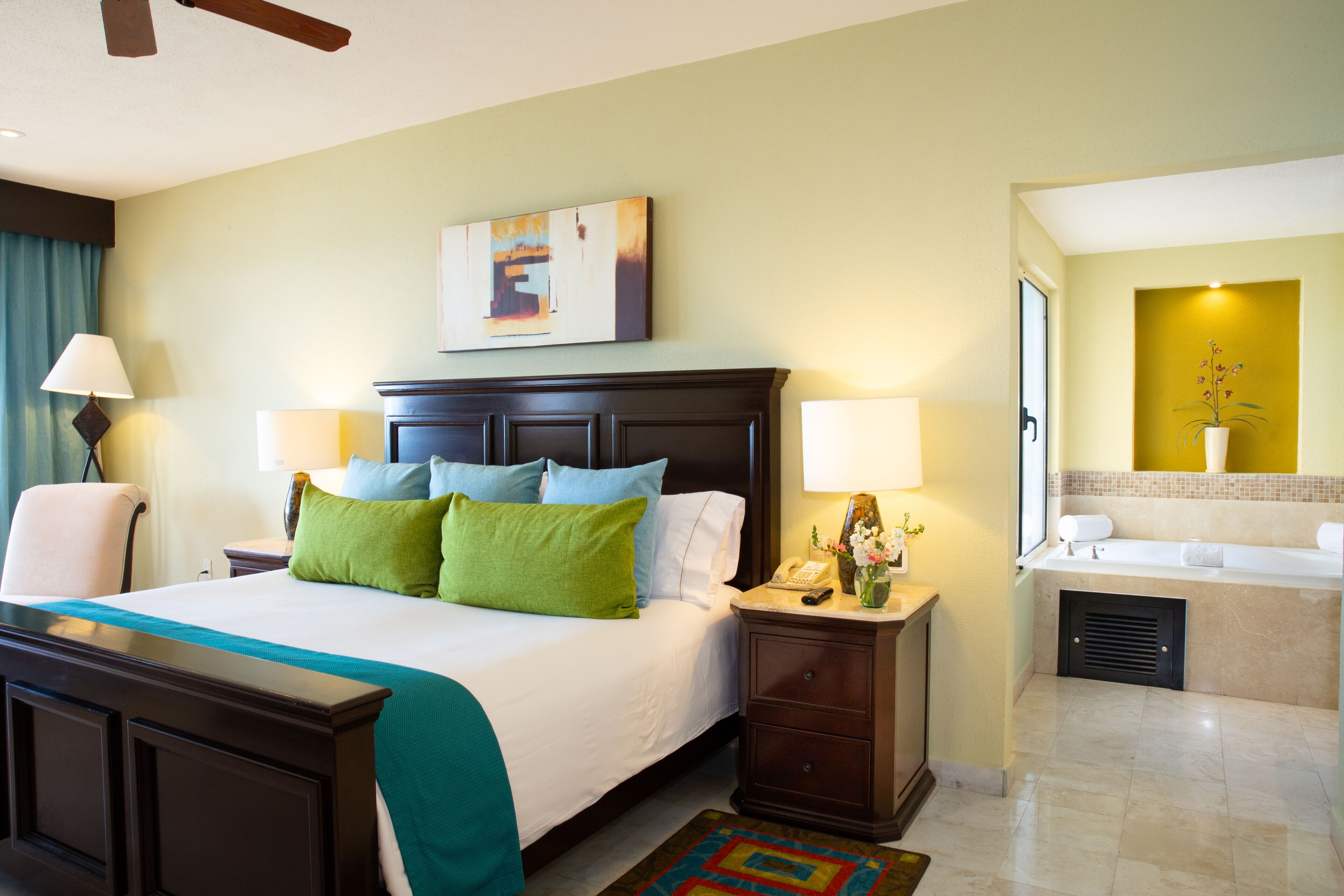 A hotel room with a large bed, bedside tables, a desk, a chair, a bathroom with a bathtub, and a ceiling fan.