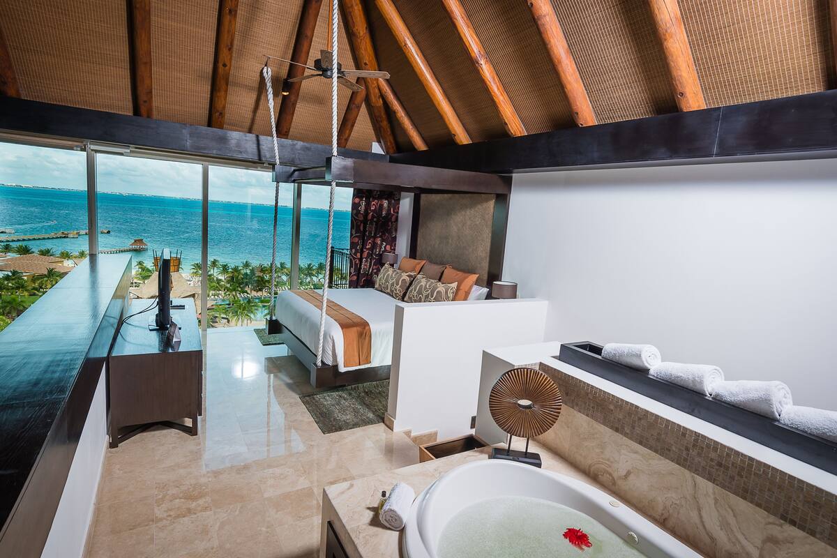 A modern bedroom with a large window overlooking the ocean, a private balcony, and a spacious bathroom with a bathtub and towel rack.