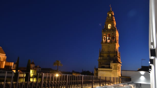 A terrace with white plastic chairs and tables, a metal railing, and a historic tower in the background. - Hotel Marisa (Córdoba)