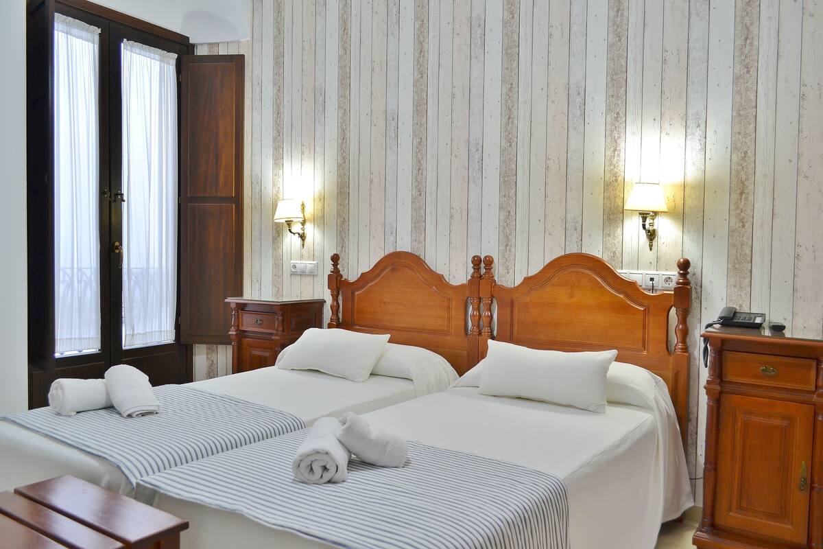 A hotel room with two beds, wooden headboards, white linens, striped bedding, a wooden nightstand, a lamp, and a wooden wardrobe.