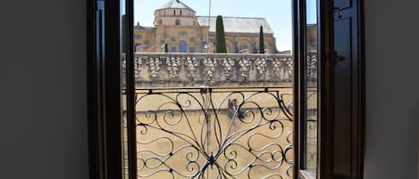Vista a través de una ventana con celosía de hierro ornamentada, un balcón y un edificio con cúpula y arcos al fondo.