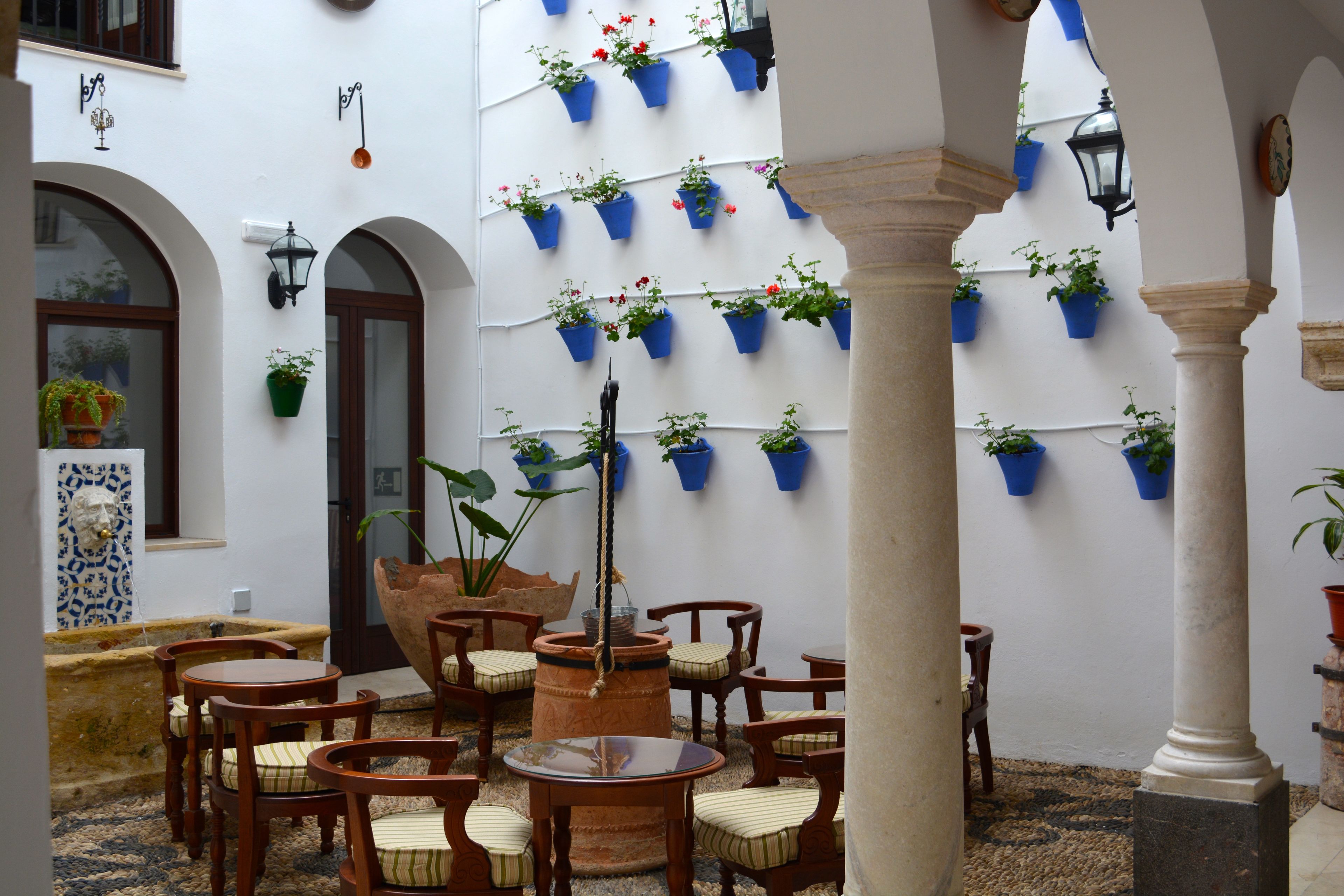 Une cour avec des tables et des chaises en bois, un grand pot en terre cuite et des jardinières bleues suspendues aux murs.