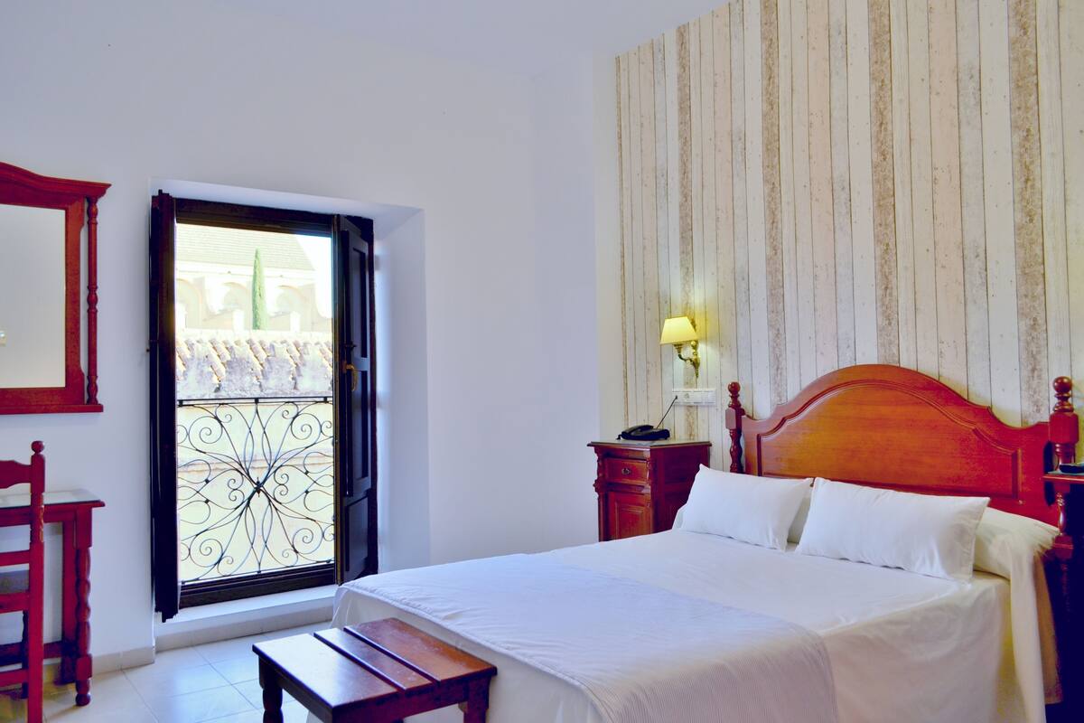 A bedroom with a bed, a wooden nightstand, a bedside table, a lamp, a mirror, and a window with a decorative iron grill.
