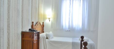 Una habitación pequeña y limpia con una cama individual, un aparador de madera y un ventanal con cortinas.