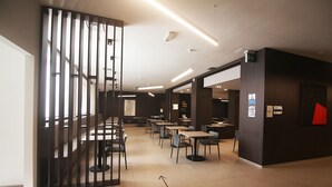 Un comedor moderno y bien iluminado, con varias mesas y sillas, un separador con listones verticales y una obra de arte geométrica roja montada en la pared.