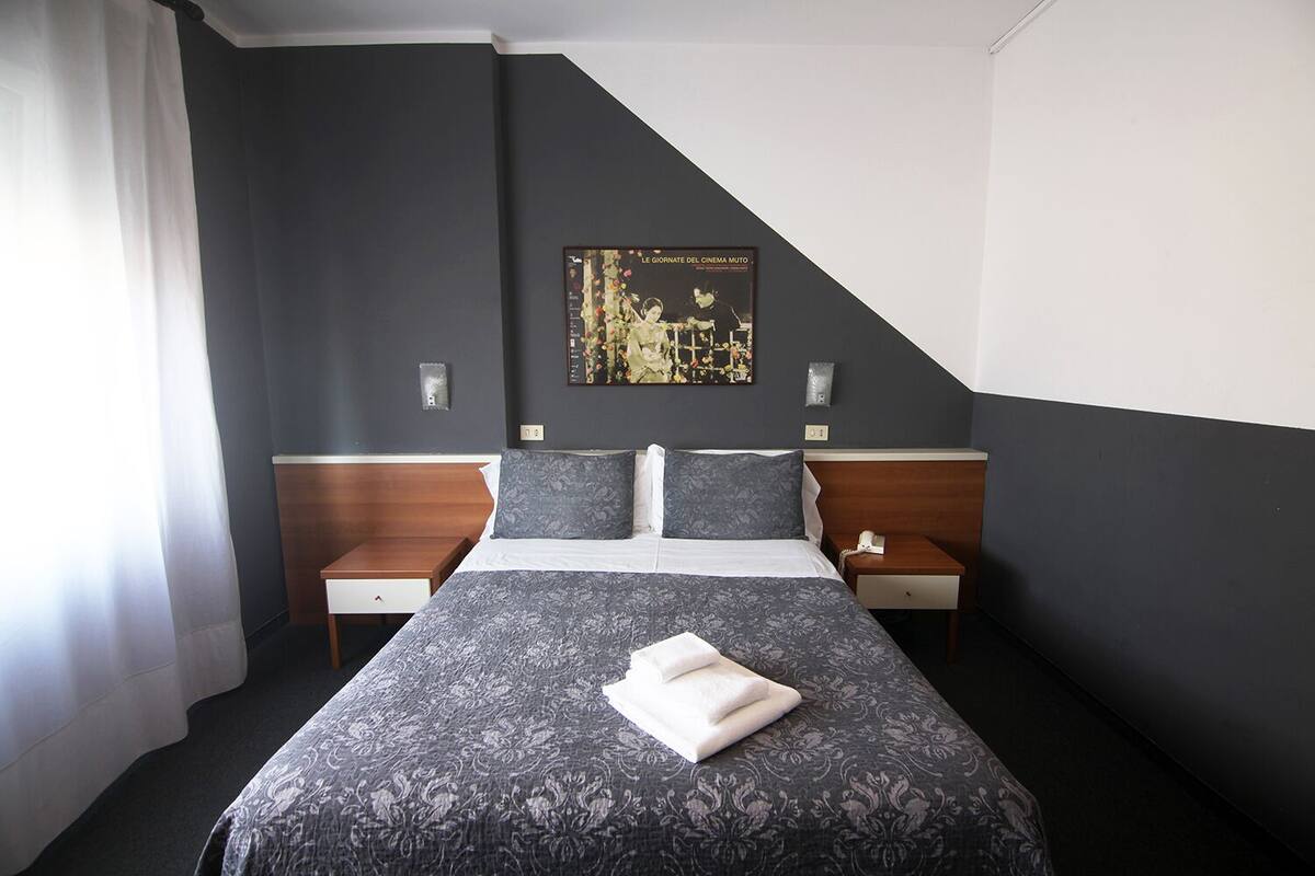 Una habitación de hotel con una cama, dos mesitas de noche, una lámpara de pared e imagen en un marco colgado en la pared.