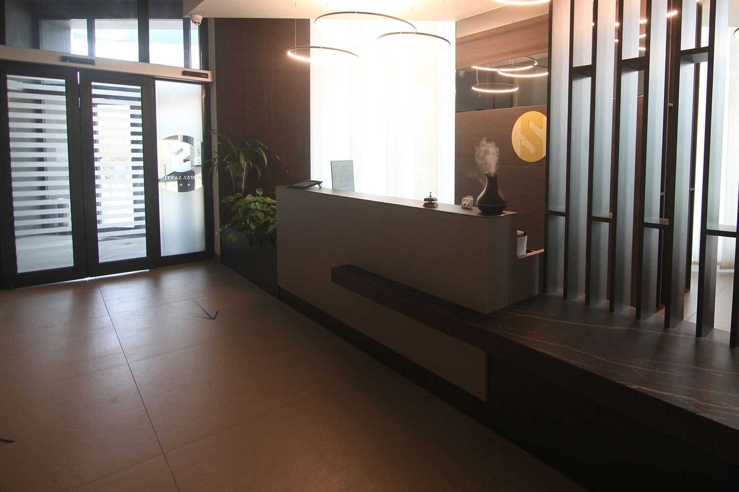 Et receptionsområde med receptionsdisk, en potteplante og et stort vindue, der lukker naturligt lys ind.