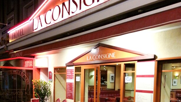Un espace extérieur aménagé avec des chaises et des tables en osier, une enseigne indiquant « BAR LA CONSIGNE » et un panneau affichant le menu, visible depuis l’entrée.