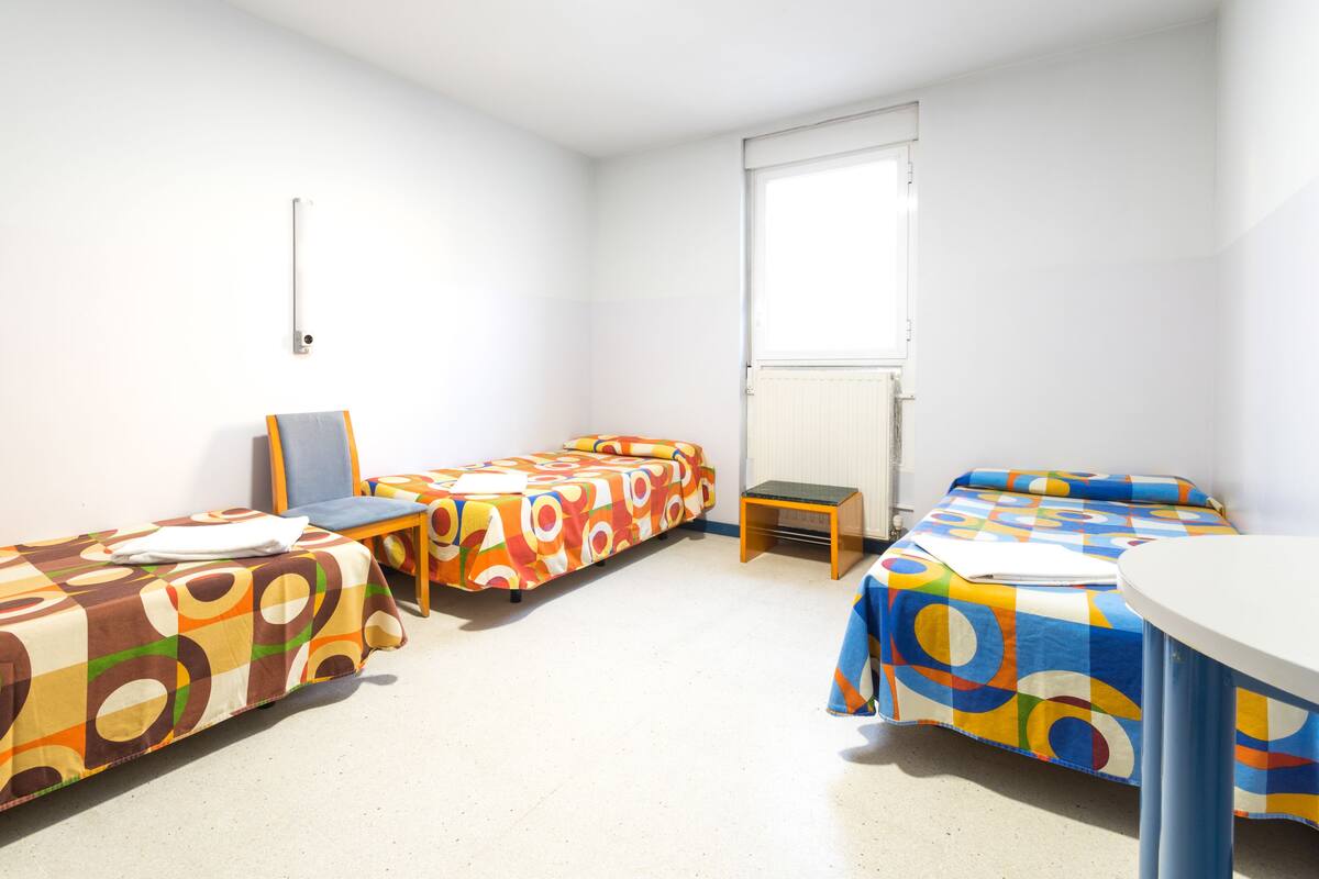Quarto de estudante com duas camas de solteiro, uma cadeira, uma mesa e uma janela.
