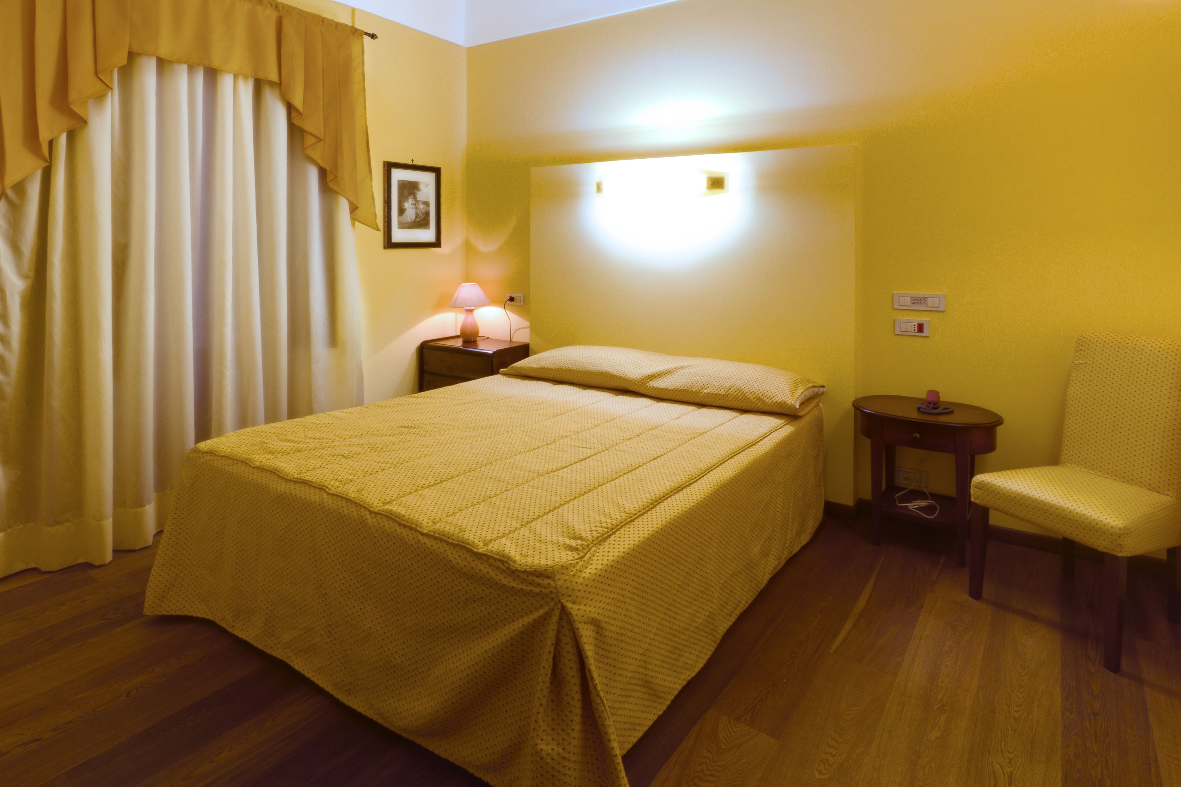 Una habitación de hotel con cama, una silla, una mesita de noche con lámpara y una luz en la pared.