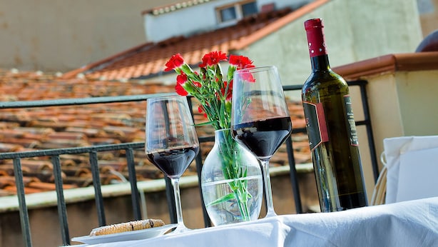 A table with a bottle of wine, two wine glasses, and a vase with flowers on a balcony overlooking a historic building with towers. - La Plumeria Hotel (Cefalù)