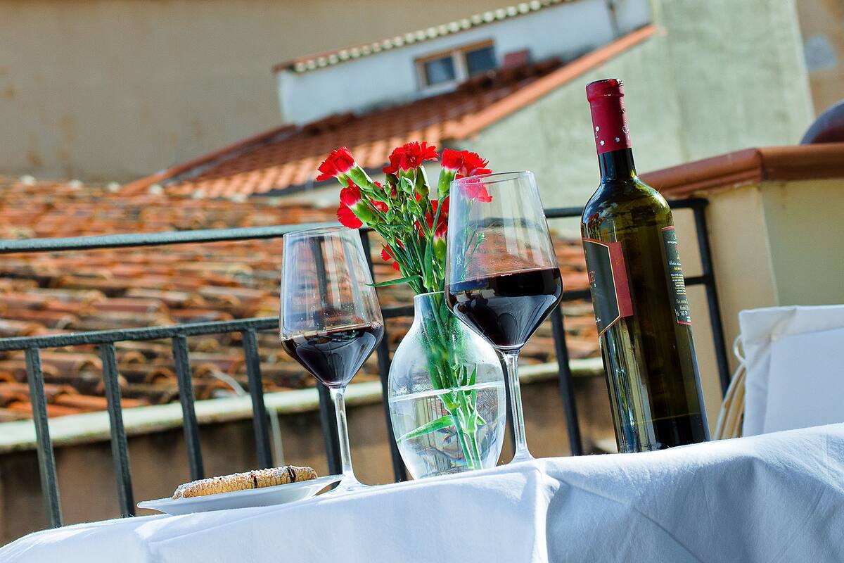 Tafel met een fles wijn, twee wijnglazen en een vaas bloemen op een balkon met uitzicht op een historisch gebouw met torens.