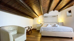 A bedroom with a wooden ceiling, a bed with white linens, a white armchair, and a bedside table with a lamp. - La Plumeria Hotel (Cefalù)