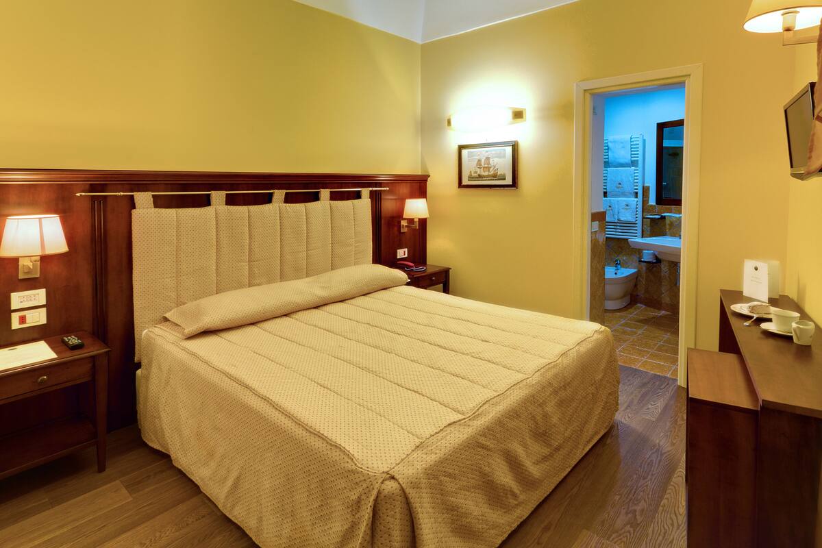 Hotelkamer met een bed, nachtlampen, een nachtkastje, een bureau met stoel, en een zichtbare badkamer door een open deur.