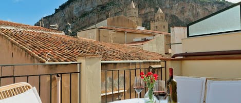 Una terraza con sillas de mimbre y una mesa lista para comer, con vistas a un edificio histórico y un paisaje montañoso.
