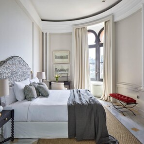 Quarto com uma cama grande, uma escrivaninha com luminária, um banco vermelho e vista pela janela.