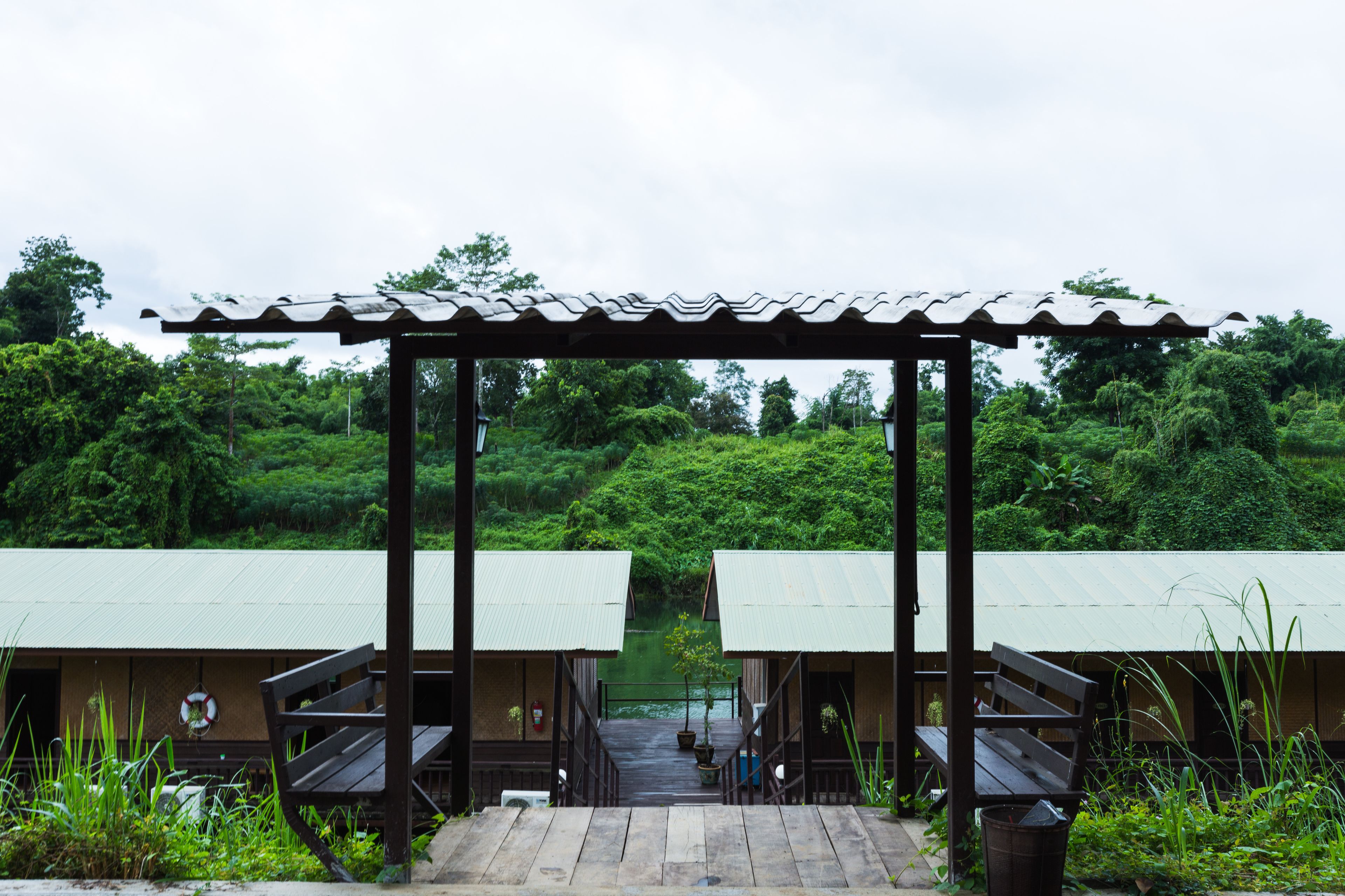 A wooden walkway leading to two small, single-story buildings with green roofs, surrounded by lush greenery.