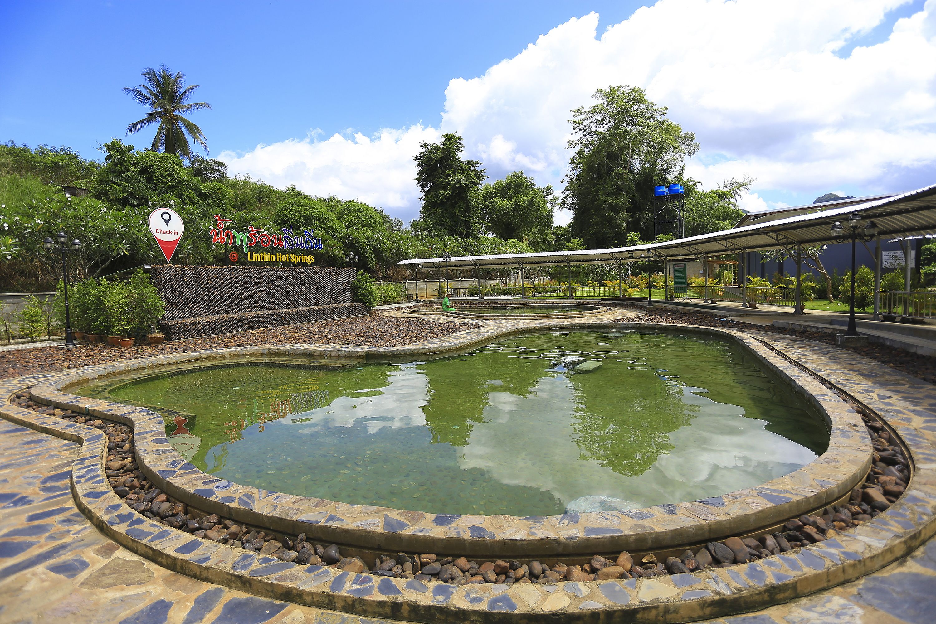 Una piscina con borde de piedra, una zona de estar techada y un cartel con texto en tailandés.