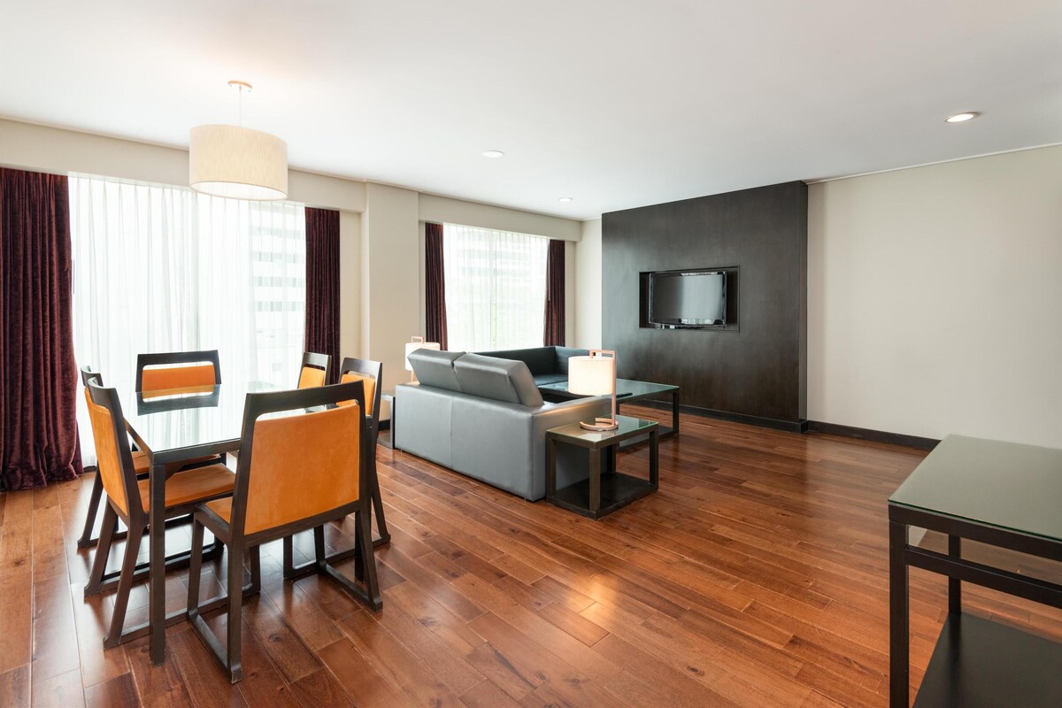 A modern hotel room with a sofa, dining table, chairs, a TV, and a large window with curtains.