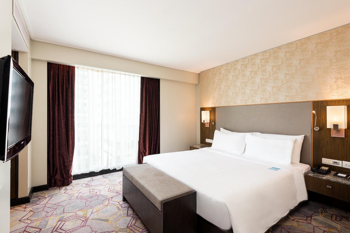 Ein Hotelzimmer mit einem großen Bett, einem Flachbildfernseher, einem Nachttisch mit Lampe und einem Fenster mit Vorhängen.