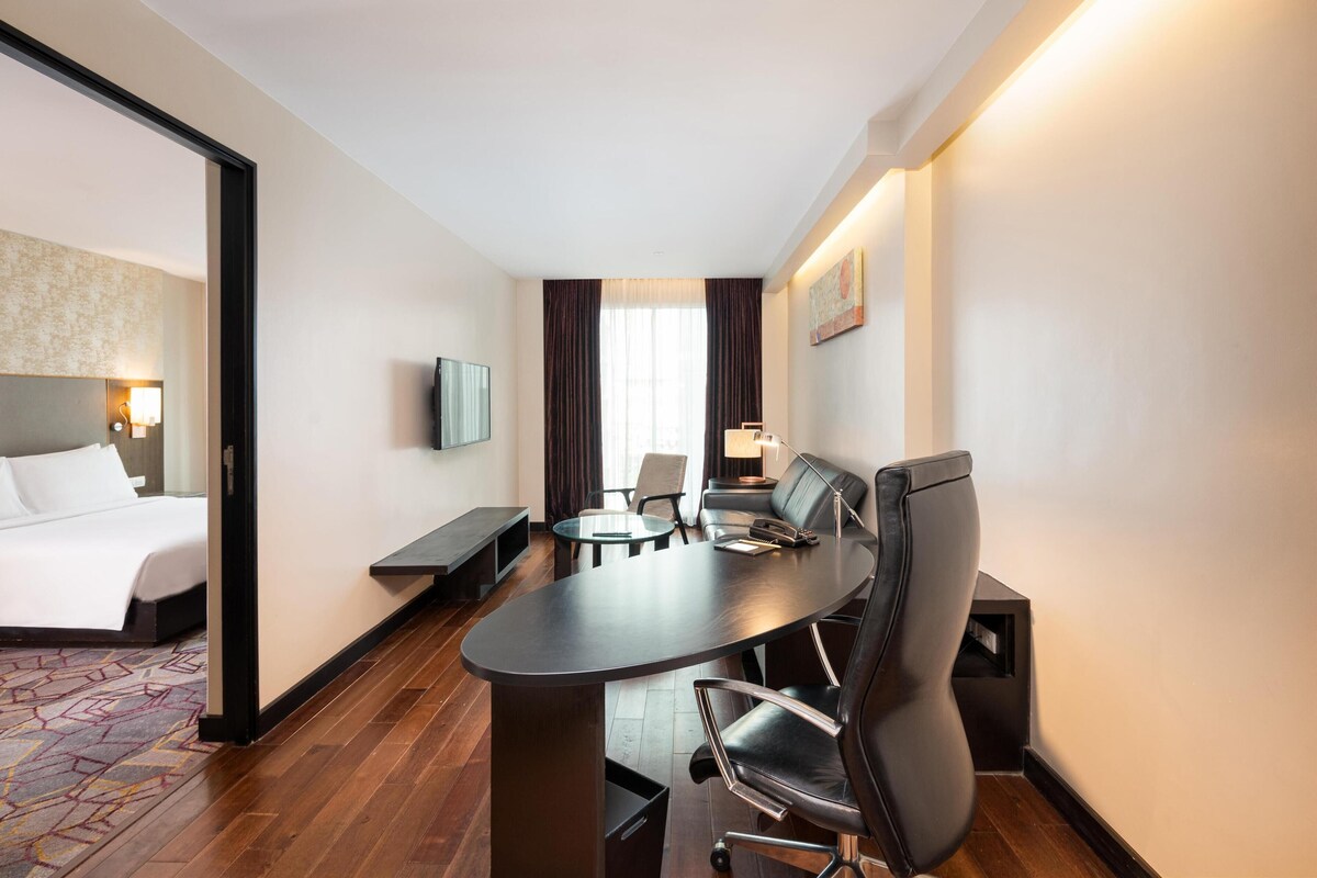 A hotel room with a bed, a dining table with chairs, a TV, and a window with curtains.