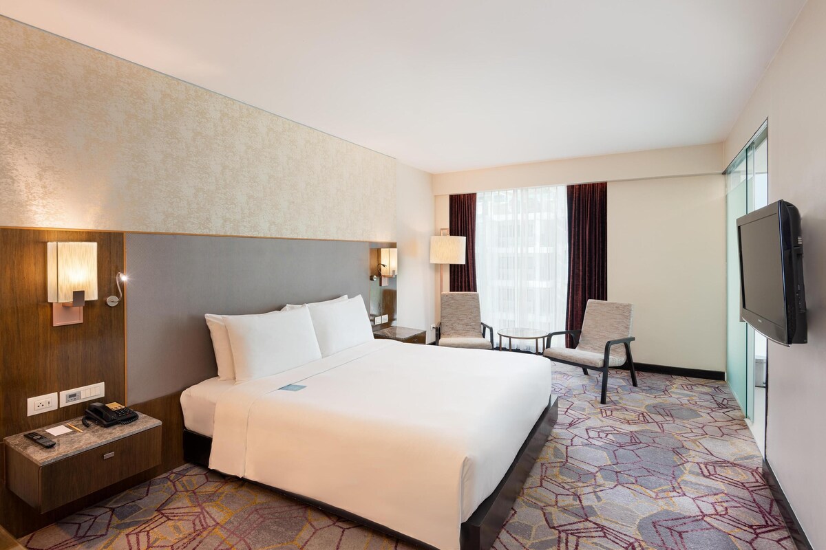 A hotel room with a large bed, a desk with a telephone, a chair, a TV, and a window with curtains.