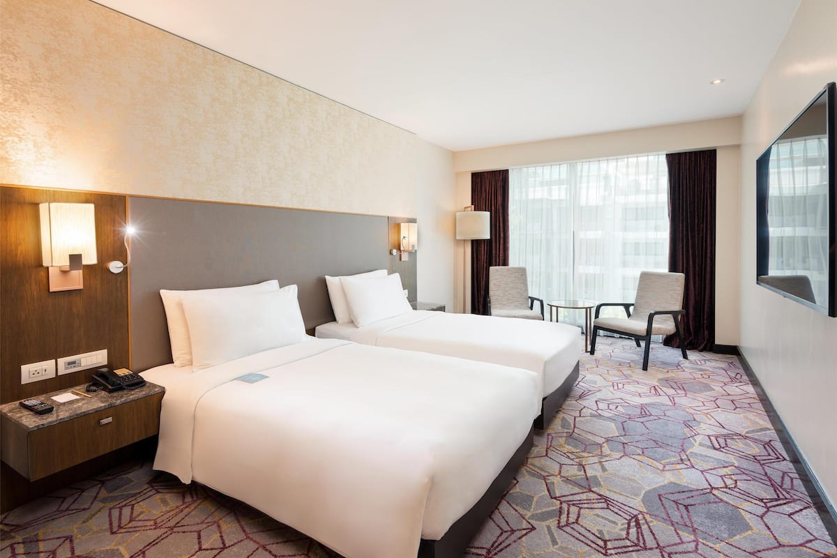 Ein Hotelzimmer mit zwei Betten, einem Schreibtisch mit Telefon, einem Sessel, einem großen Fenster mit Vorhängen und einem am Wand montierten Flachbildfernseher.