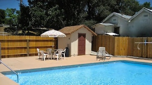 Una piscina con cerca de madera, un pequeño edificio con puerta roja y una mesa con sillas.