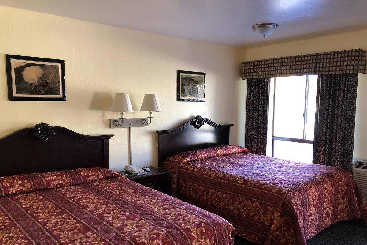 Een hotelkamer met twee bedden, een nachtkastje met telefoon en lamp, en een raam met gordijnen.