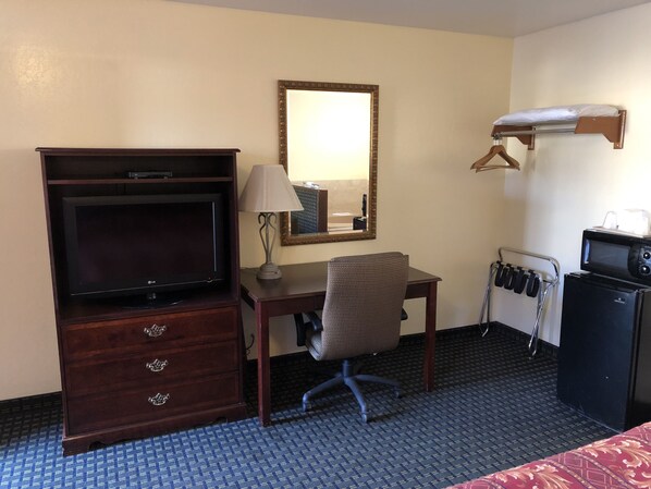 A hotel room with a wooden dresser, a desk with a chair, a television, a microwave, and a mirror on the wall.