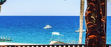 Una terraza de madera con mesas y sillas que da a un mar azul y transparente, con barcos a lo lejos.