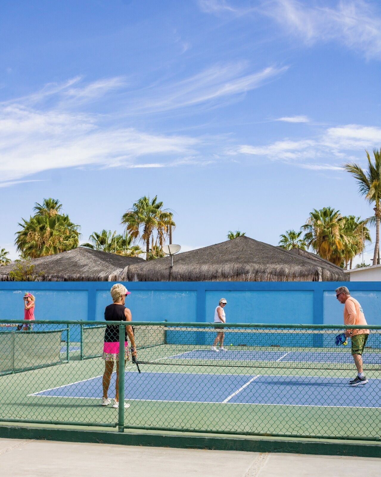Una cancha de tenis con jugadores, rodeada de palmeras y un muro azul.