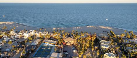 Vista aérea de un complejo turístico costero con playa, canchas de tenis y palmeras.
