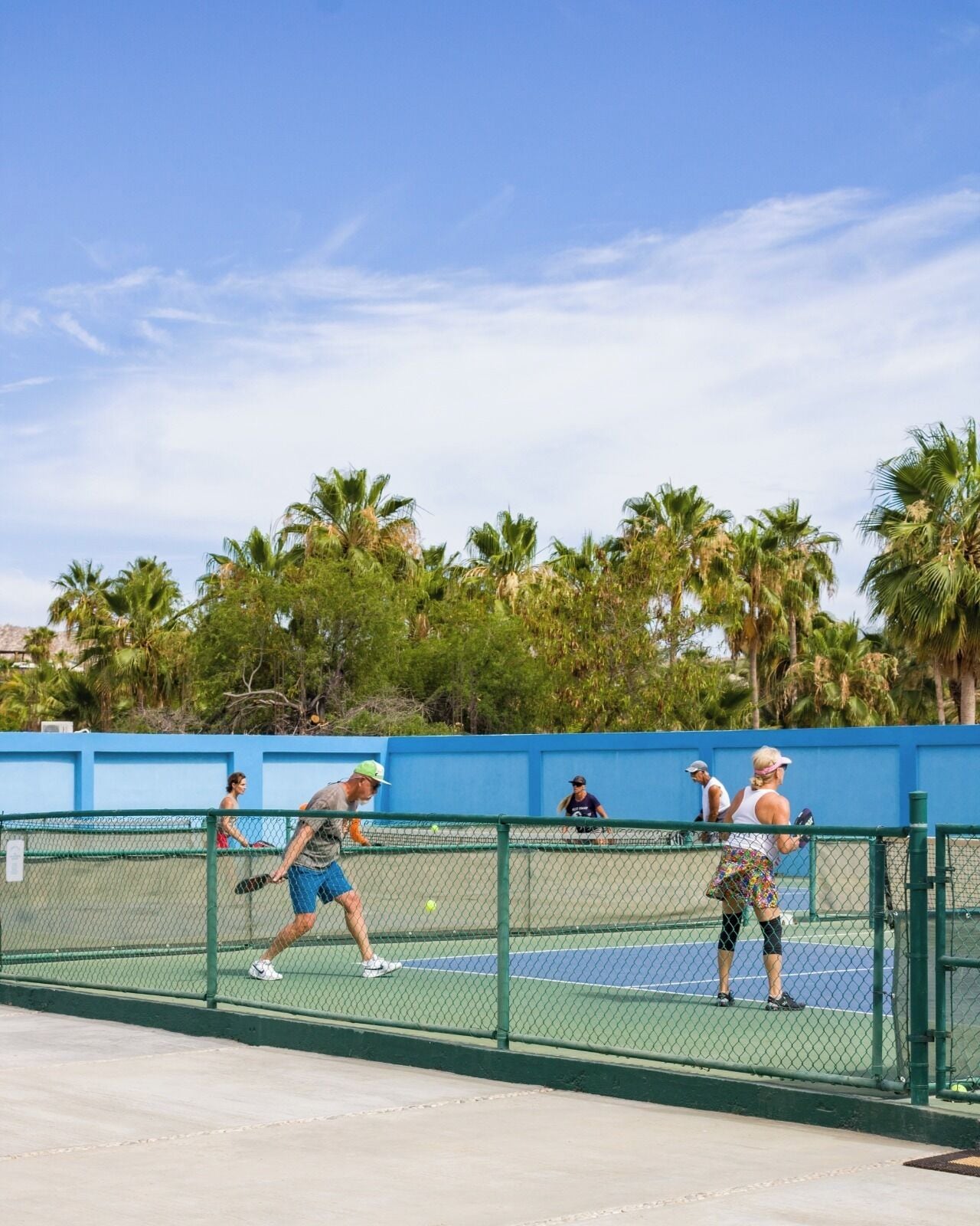 Ein Tennisplatz mit Spielern, umgeben von einem blauen Zaun und Palmen.