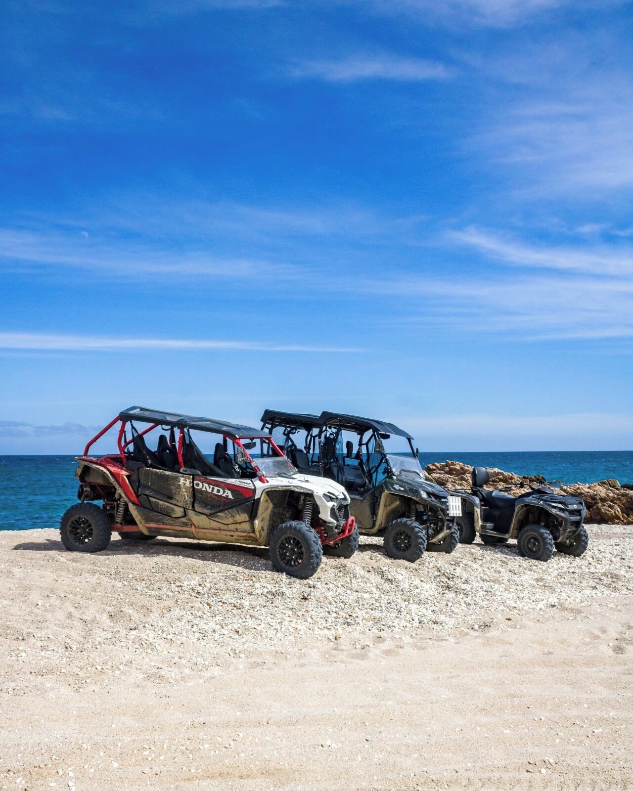 Three off-road vehicles parked on a rocky beach with the ocean in the background.