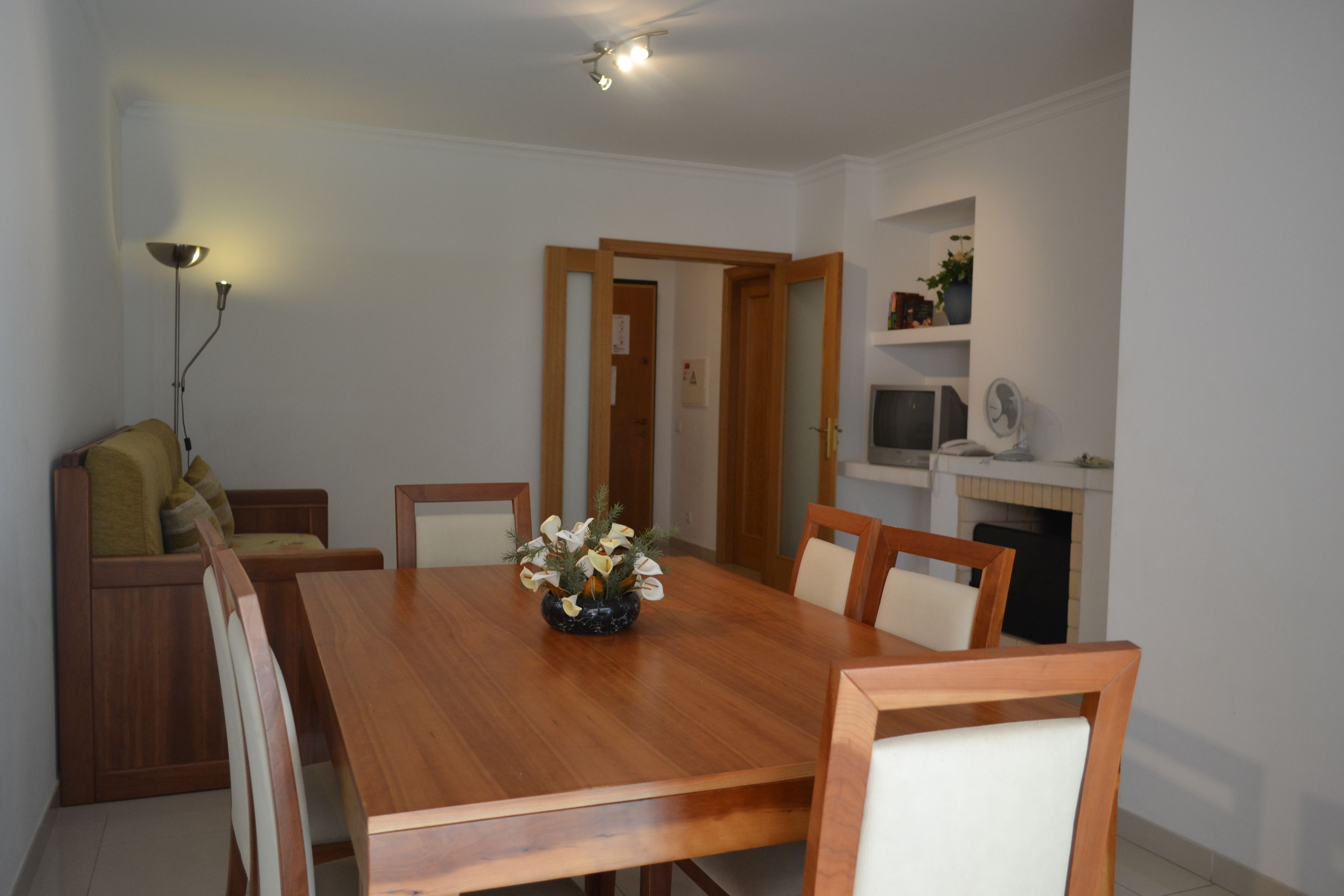A dining area with a wooden table, chairs, and a vase of flowers. There is a sofa with cushions and a lamp on the left. A television and a shelf with decorative items are on the right wall.